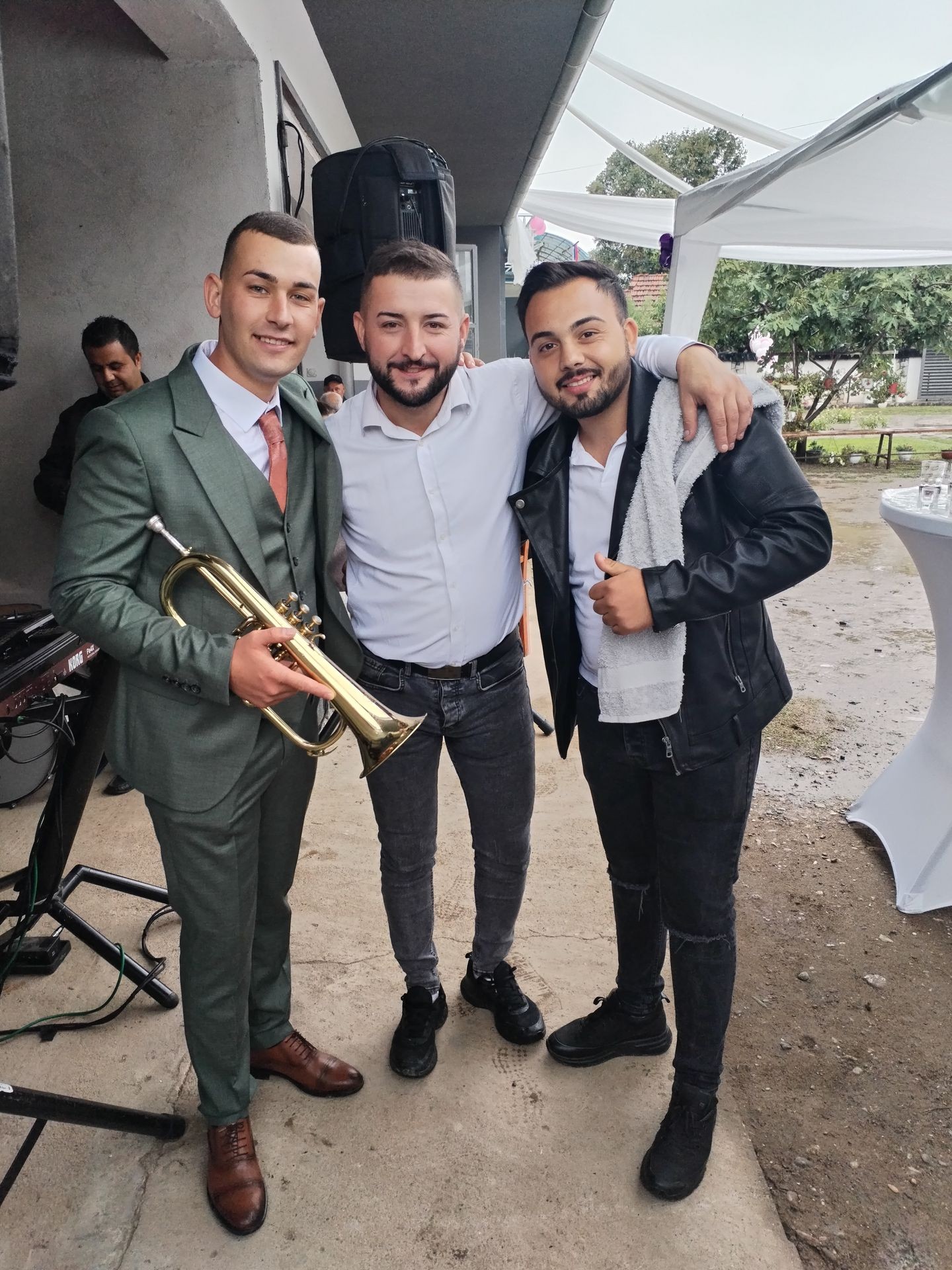 Three men posing together outdoors, one holding a trumpet, smiling at the camera.