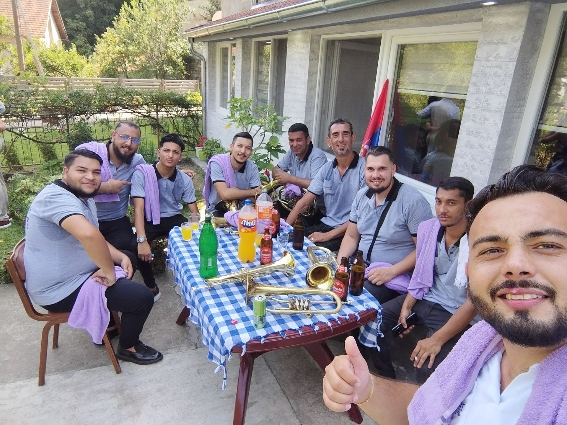 Group of men with brass instruments sitting around a table outdoors, smiling with beverages and snacks.